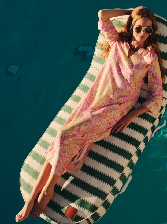 Overhead view of a woman in a colorful patterned caftan and sunglasses lounging on a striped pool float.
