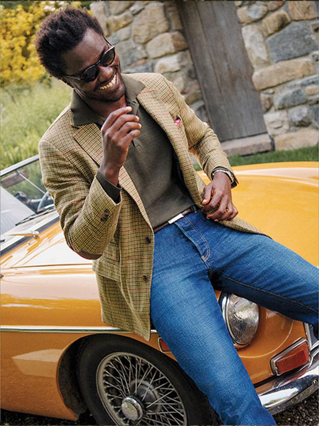 Male model sitting on a car wearing a brown sport coat and denim jeans.