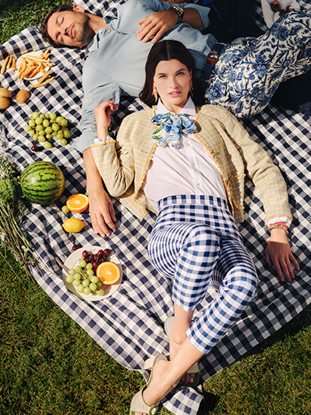 A stylishly dressed couple picnics on a blue and white checkered blanket, surrounded by fruits and fries, with the man resting and the woman looking at the camera.
