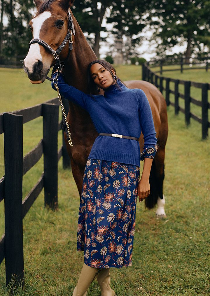 woman standing outside in a field with a horse wearing a dark blue cashmere turtleneck sweater layered over a blue floral printed dress