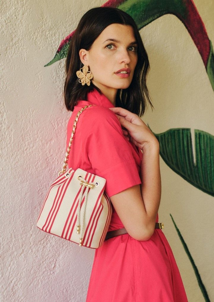 A woman in a coral dress and flower earrings holds a red and white striped bucket bag while looking over her shoulder.