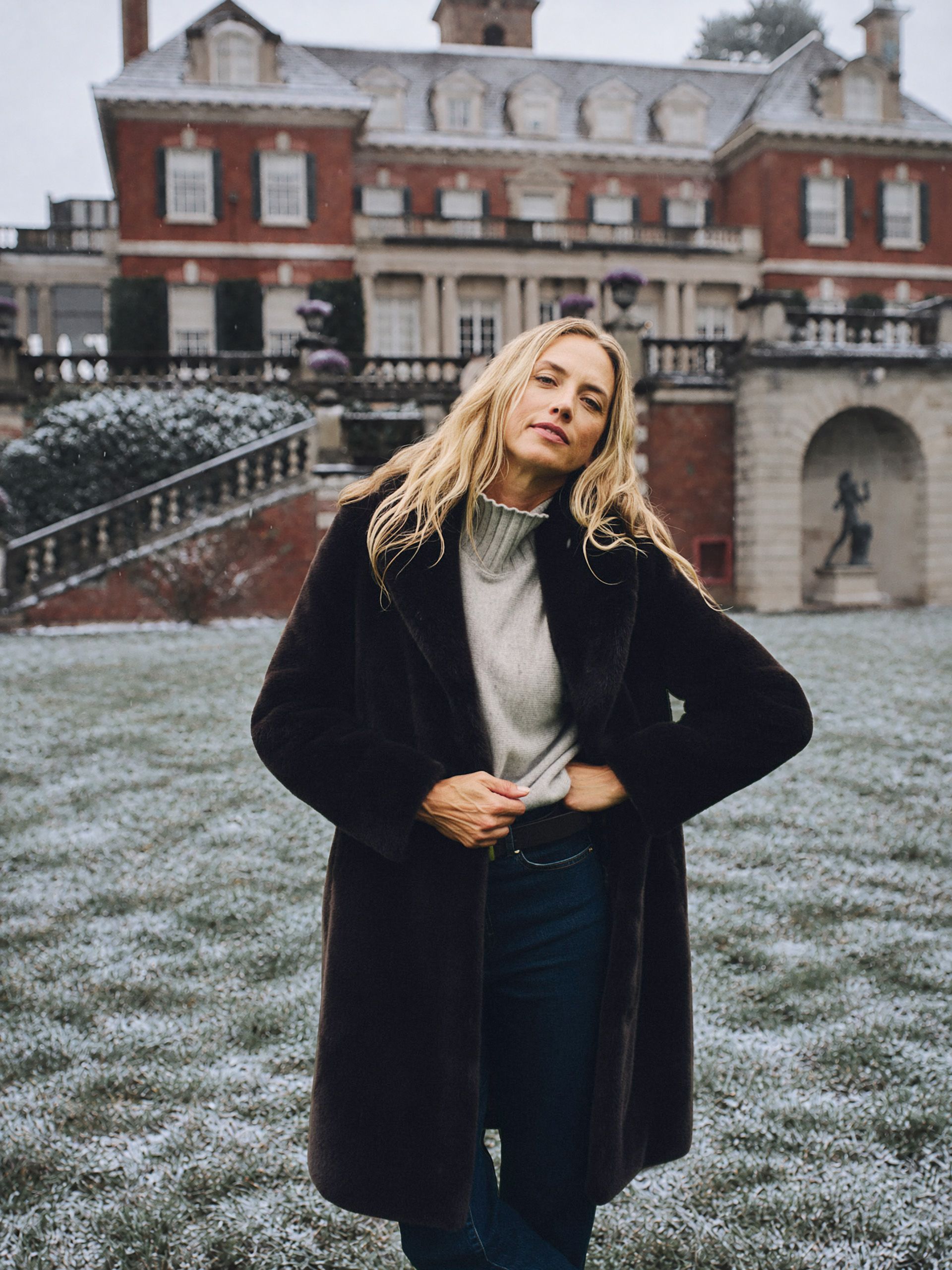 A blonde woman in a brown faux fur coat stands on snow-dusted grass in front of a grand brick mansion.