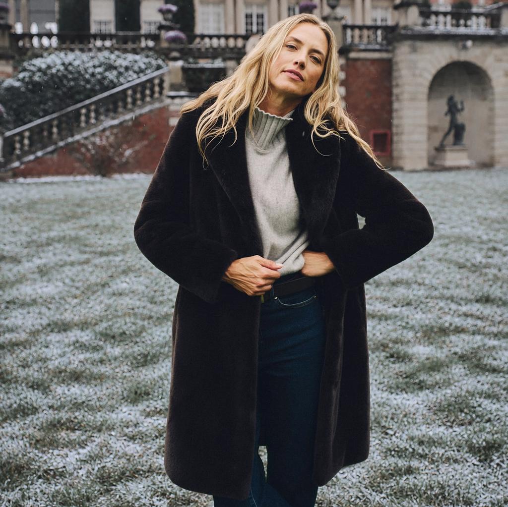 A blonde woman in a brown faux fur coat stands on snow-dusted grass in front of a grand brick mansion.