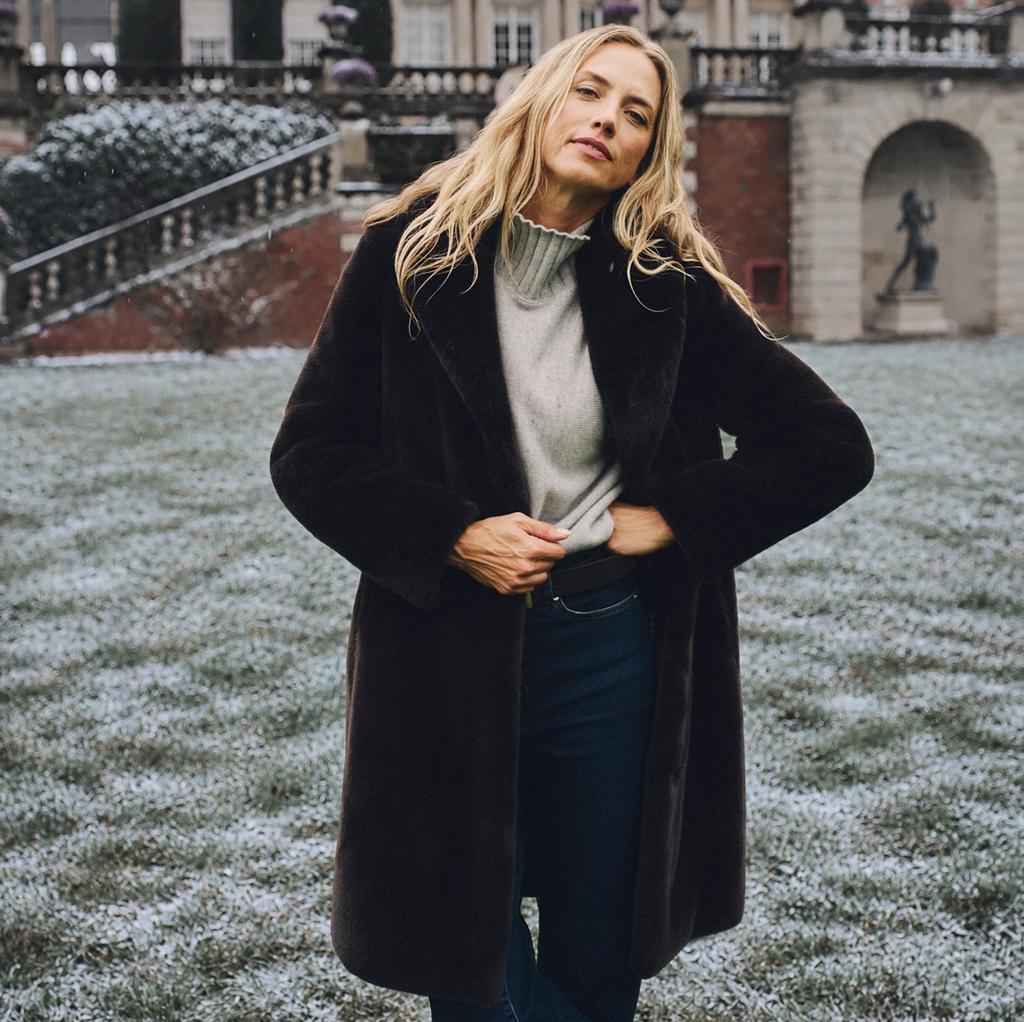 A blonde woman in a dark faux fur coat and grey turtleneck stands in a lightly snow-dusted garden in front of a grand mansion.