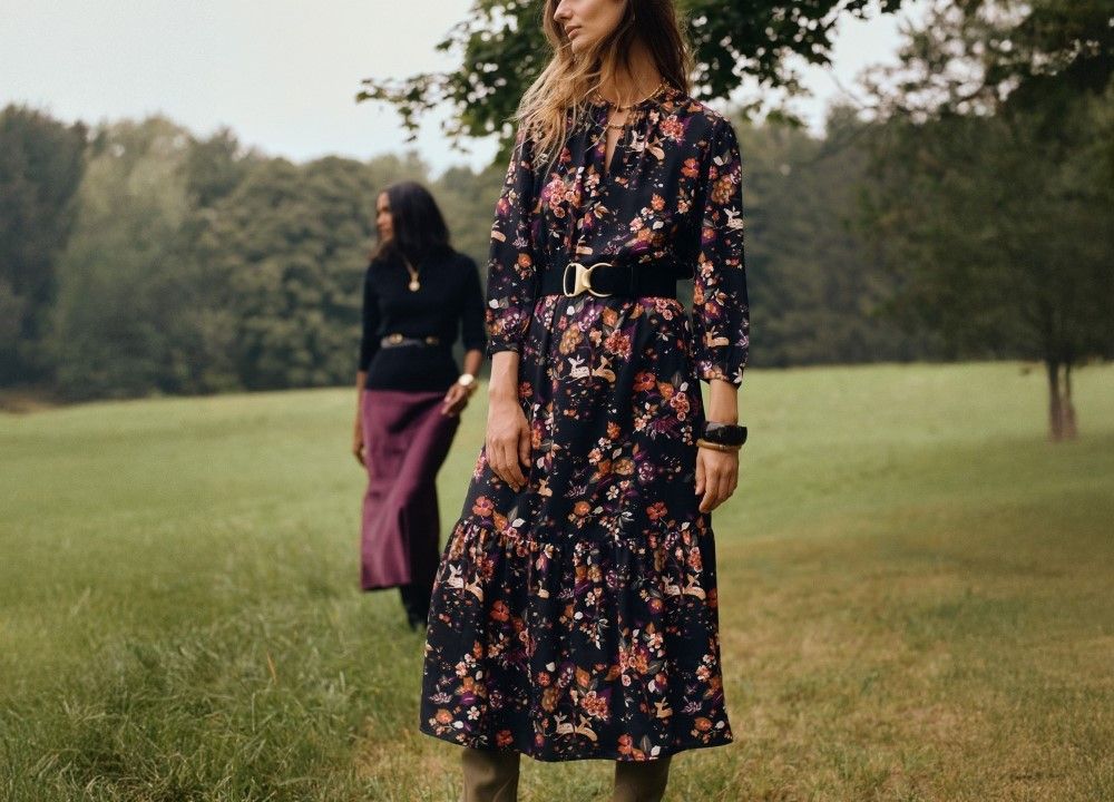 woman standing in a field wearing a black floral printed long dress, cinched with black belt at waist