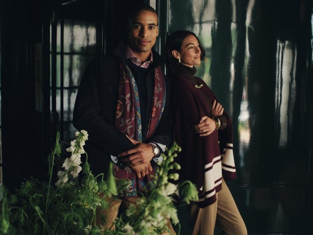 A stylish man in a dark jacket and patterned scarf stands next to a woman in a burgundy poncho with striped trim, posing against a dark green background with white flowers.