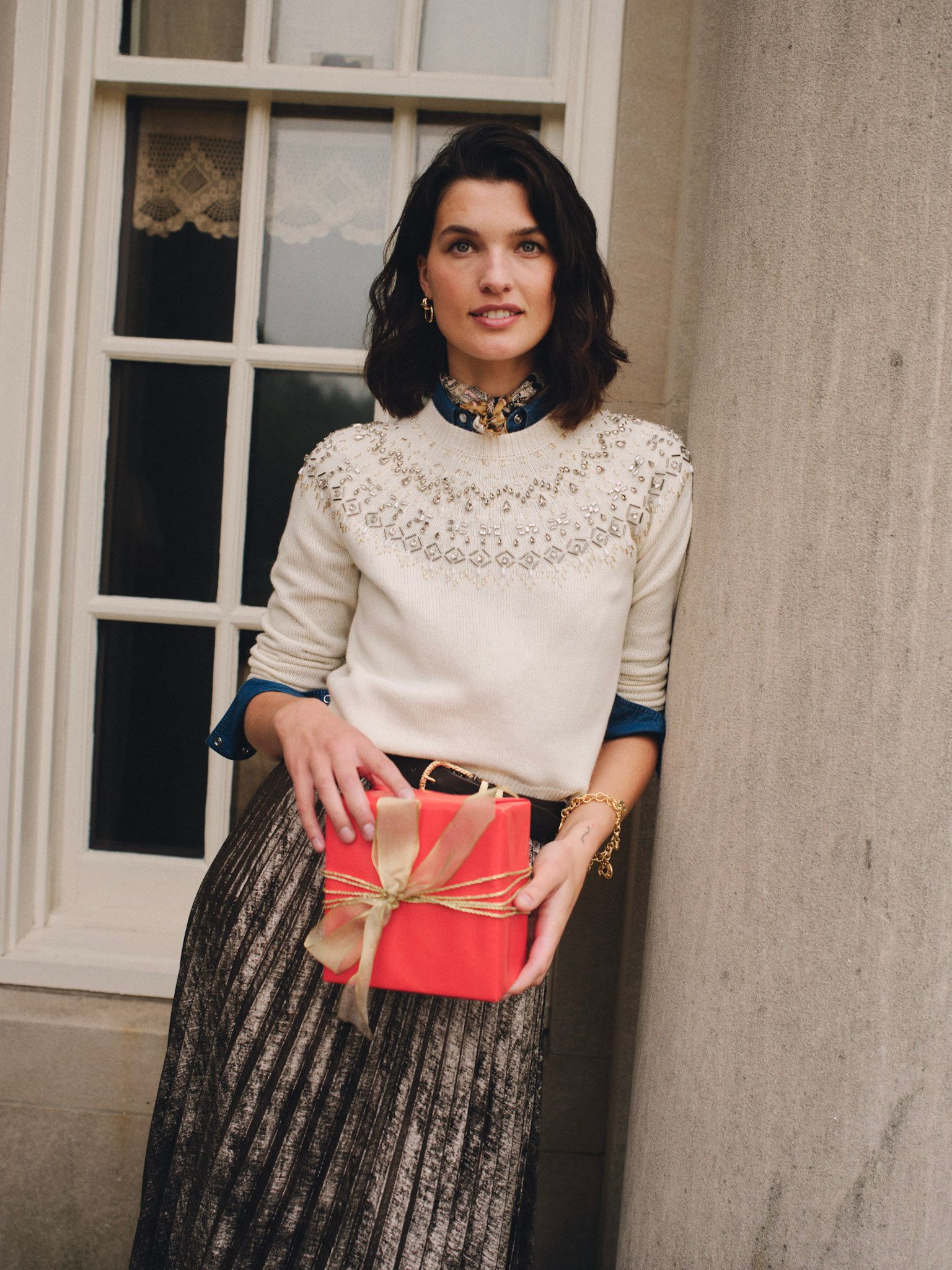 A woman in a cream beaded sweater and metallic pleated skirt holds a red gift, leaning against a stone column.