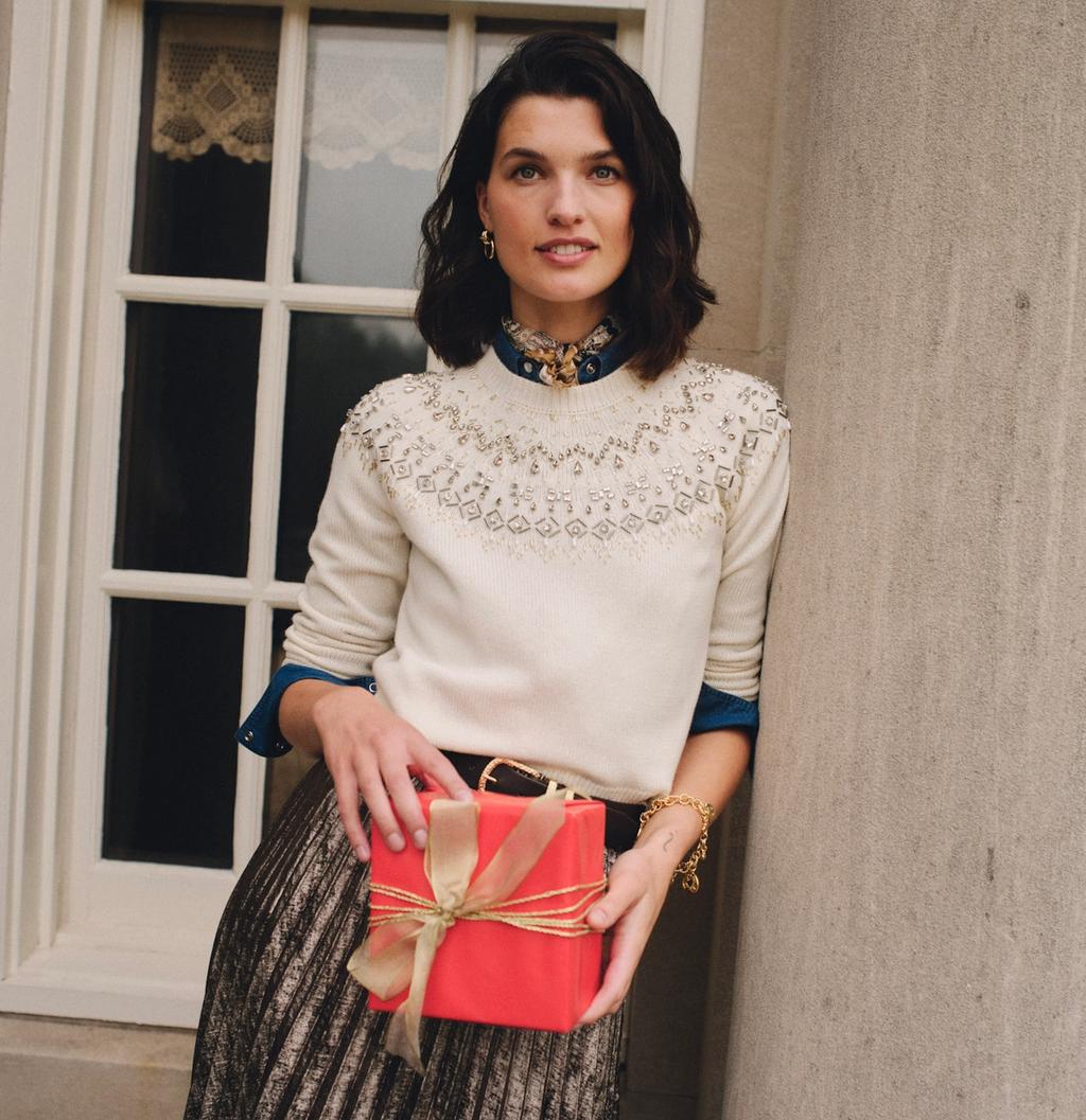 A woman in a cream beaded sweater and metallic pleated skirt holds a red gift, leaning against a stone column.