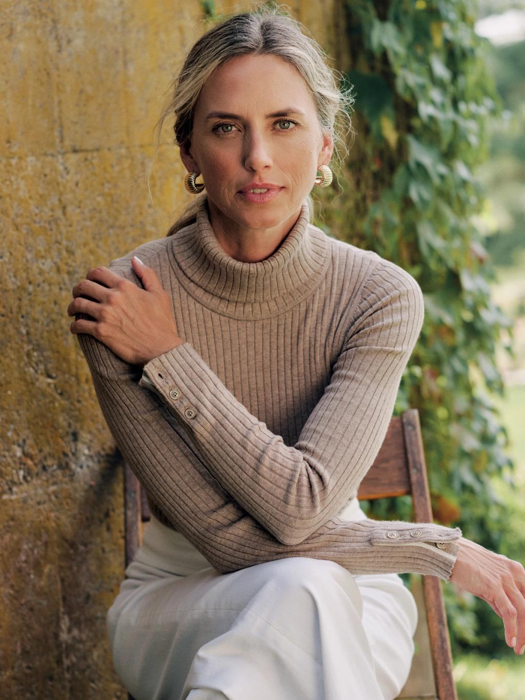 Woman with greyish-blonde hair, gold hoop earrings, beige ribbed turtleneck, and light pants sits with arms crossed, looking at camera, outdoors.