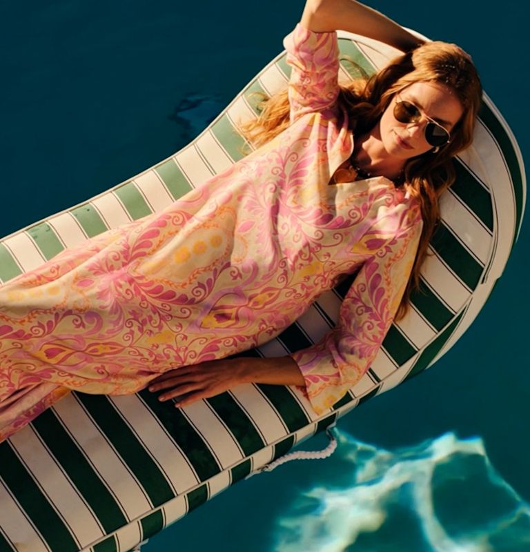A woman in a pink patterned caftan and sunglasses relaxes on a striped pool float.