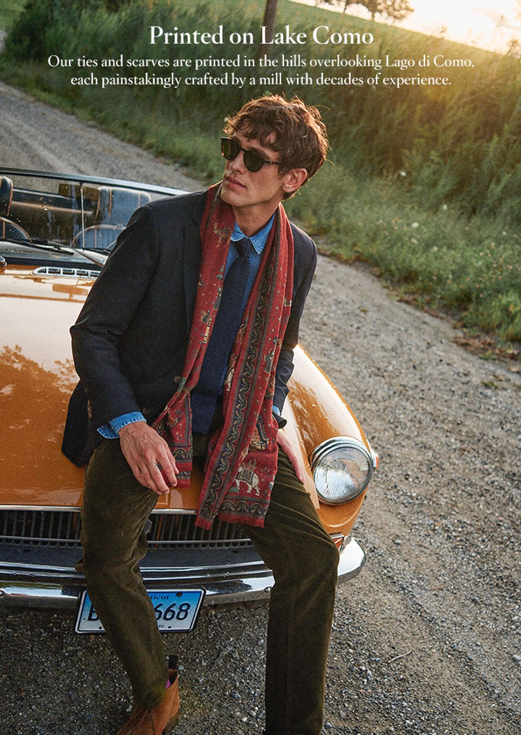 Man sitting on a car wearing corduroy pants, a navy blazer, and red scarf.