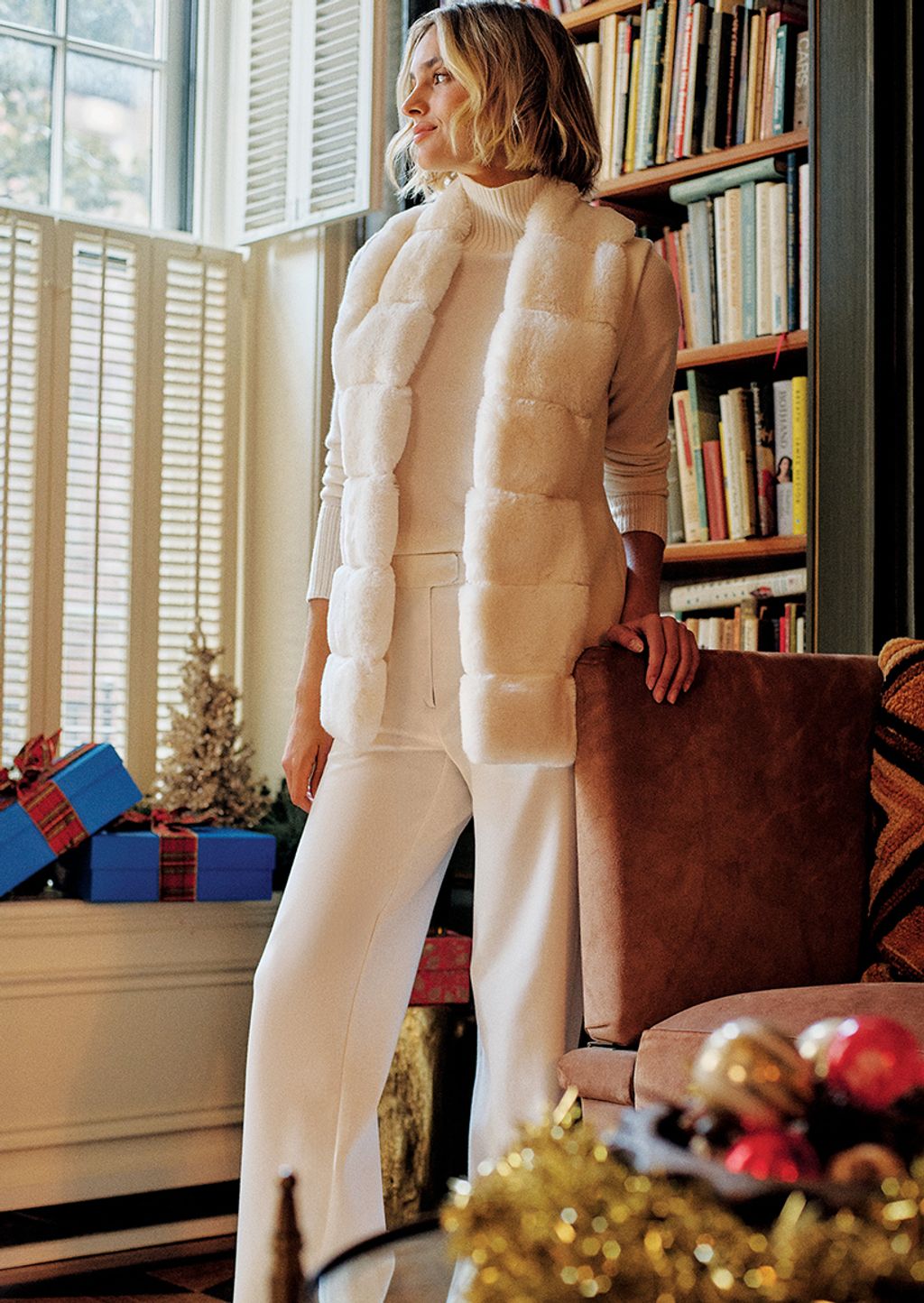 A woman in a white turtleneck sweater, white faux fur vest, and white trousers.