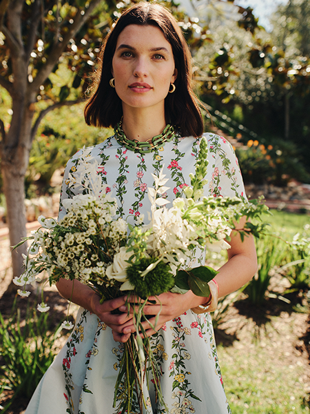 A woman stands outdoors in a garden setting, holding a lush bouquet of white and green flowers. She wears a light blue floral midi dress with pink and green botanical prints, short sleeves, and a high neckline with green trim. She has short dark hair, gold hoop earrings, and a delicate necklace. The background features soft, sun-dappled greenery and trees.