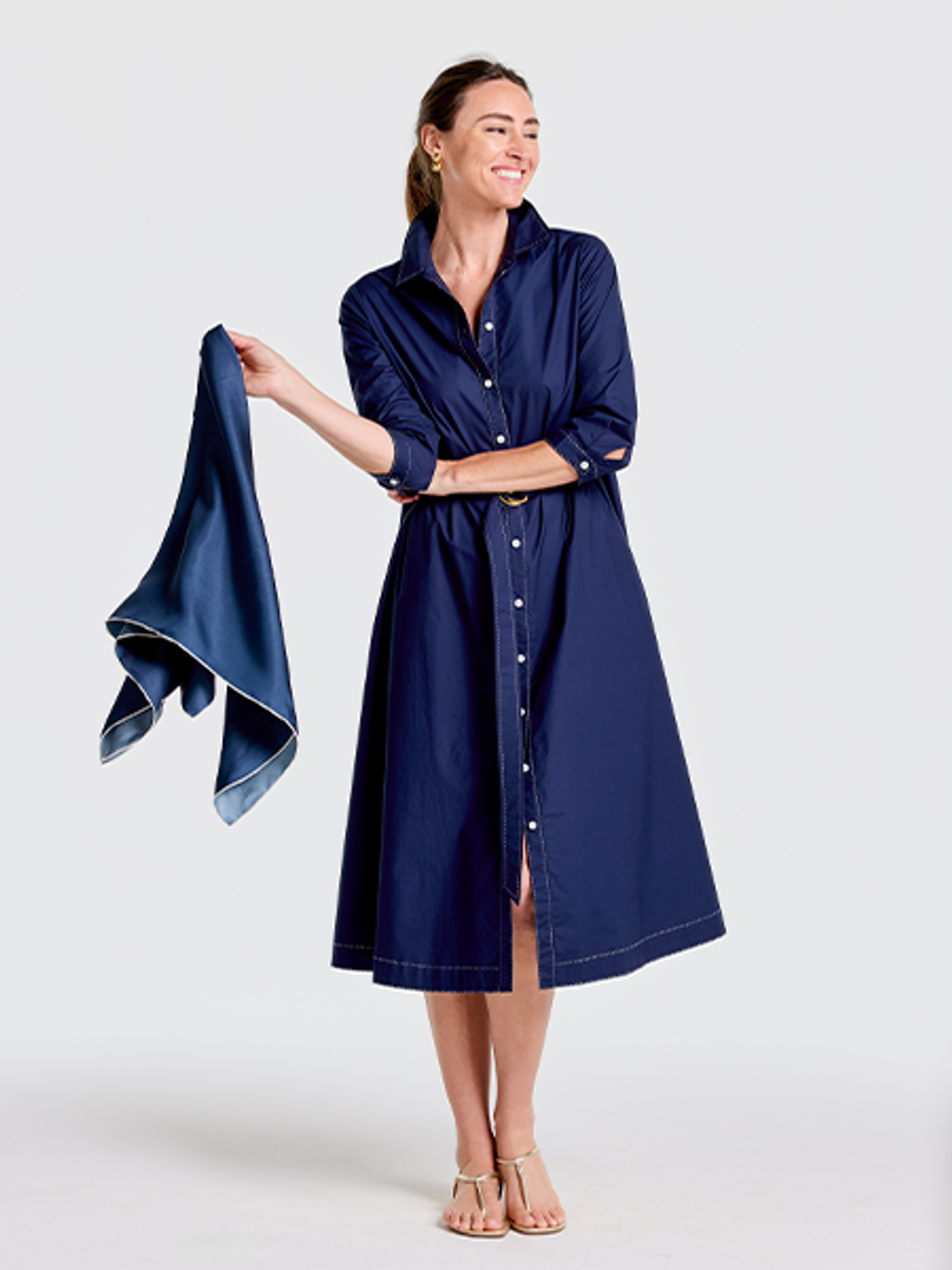 A smiling woman in a navy blue shirt dress with white stitching, holding a matching silk scarf, and wearing gold sandals.