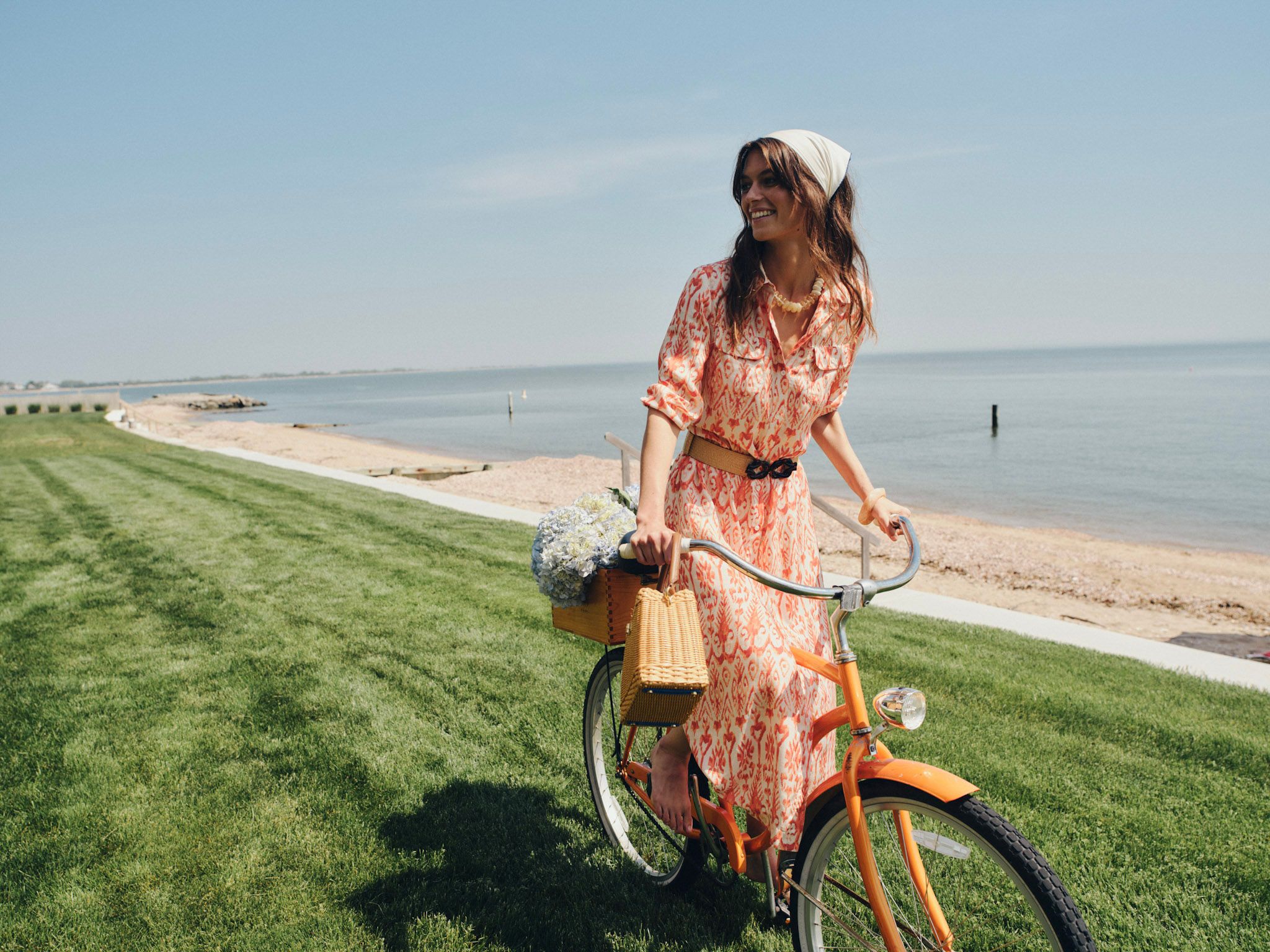 A smiling woman in a patterned dress and headscarf stands with an orange bicycle on a grassy lawn next to a sunny beach.