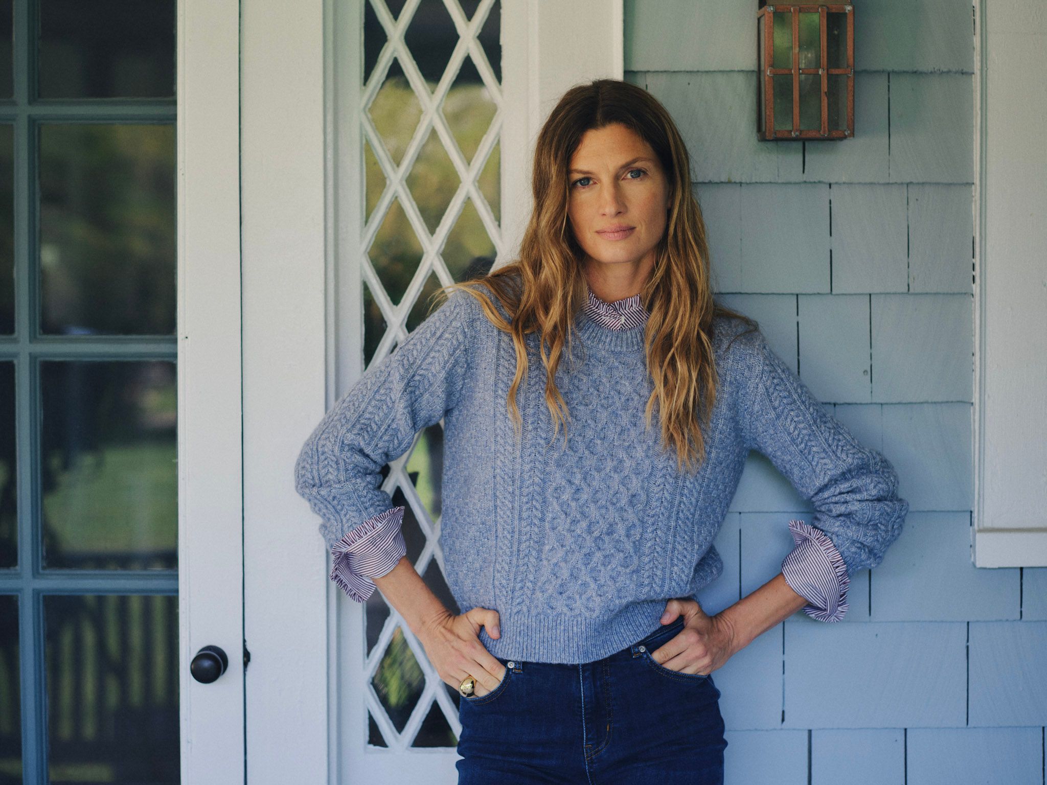 A woman with long wavy hair wears a blue cable-knit sweater and jeans, hands on hips, leaning against a light blue shingled house.