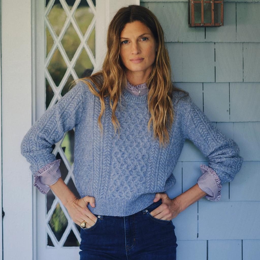 A woman in a blue cable-knit sweater and dark jeans stands with hands on hips against a blue shingled wall.