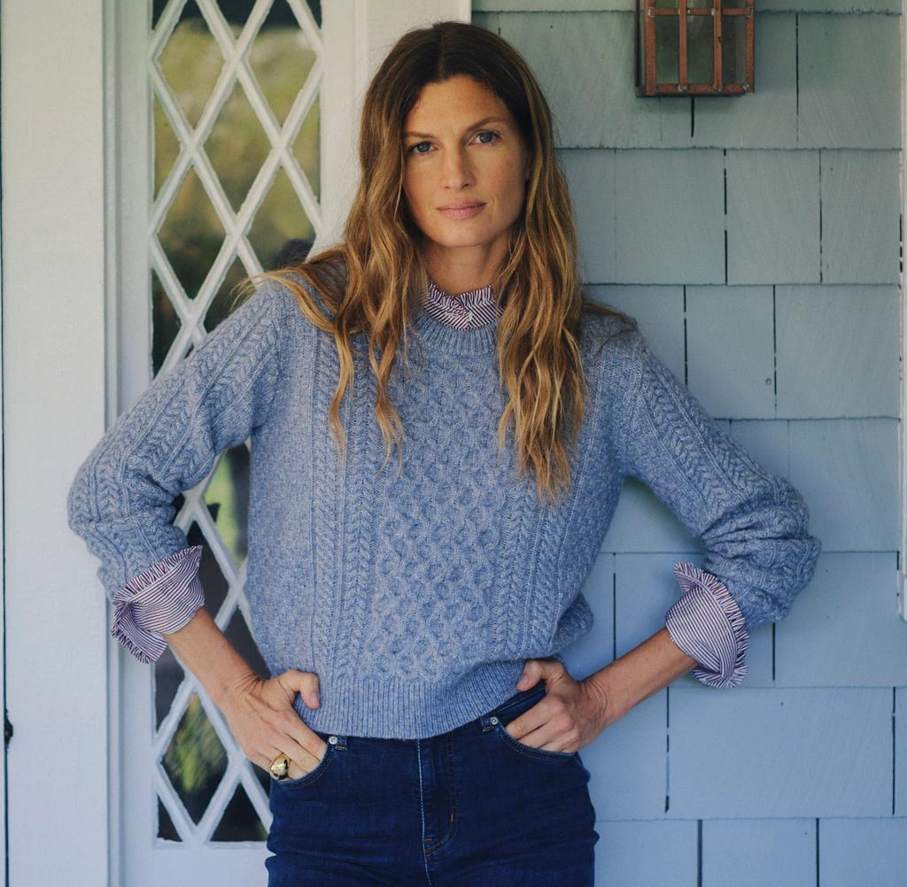 A woman with long wavy hair wears a blue cable-knit sweater and jeans, hands on hips, leaning against a light blue shingled house.