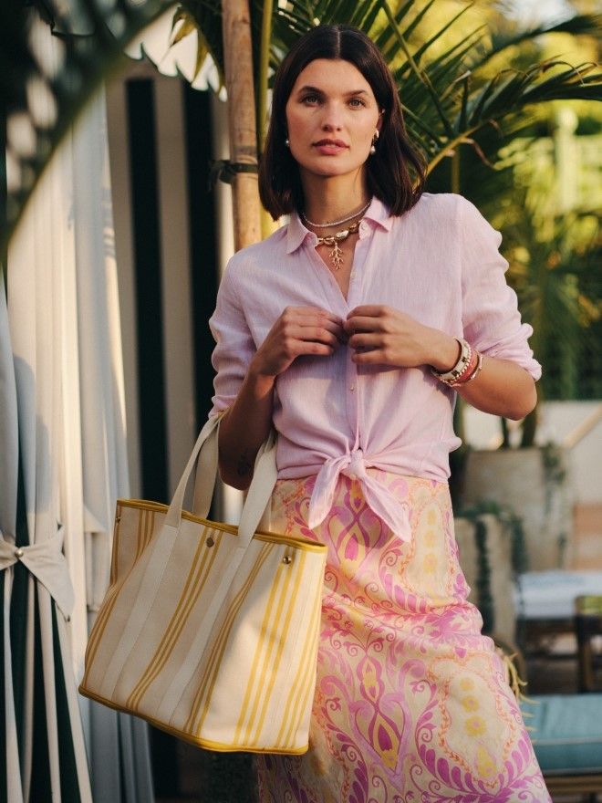 A woman wearing a pink linen shirt tied at the waist, a pink and yellow patterned skirt, and holding a cream and yellow striped tote bag in a tropical outdoor setting.