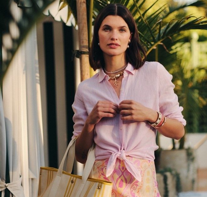A woman wearing a pink linen shirt tied at the waist, a pink and yellow patterned skirt, and holding a cream and yellow striped tote bag in a tropical outdoor setting.