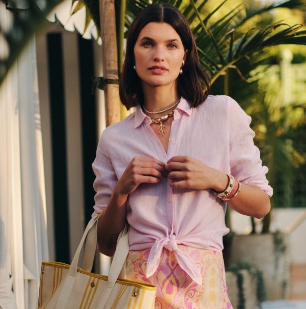 A woman wearing a pink linen shirt tied at the waist, a pink and yellow patterned skirt, and holding a cream and yellow striped tote bag in a tropical outdoor setting.