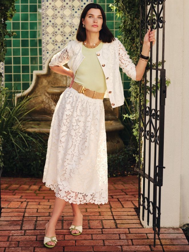 A woman wearing a light green top, white lace skirt, and white lace cardigan stands on a brick patio holding an ornate black gate.