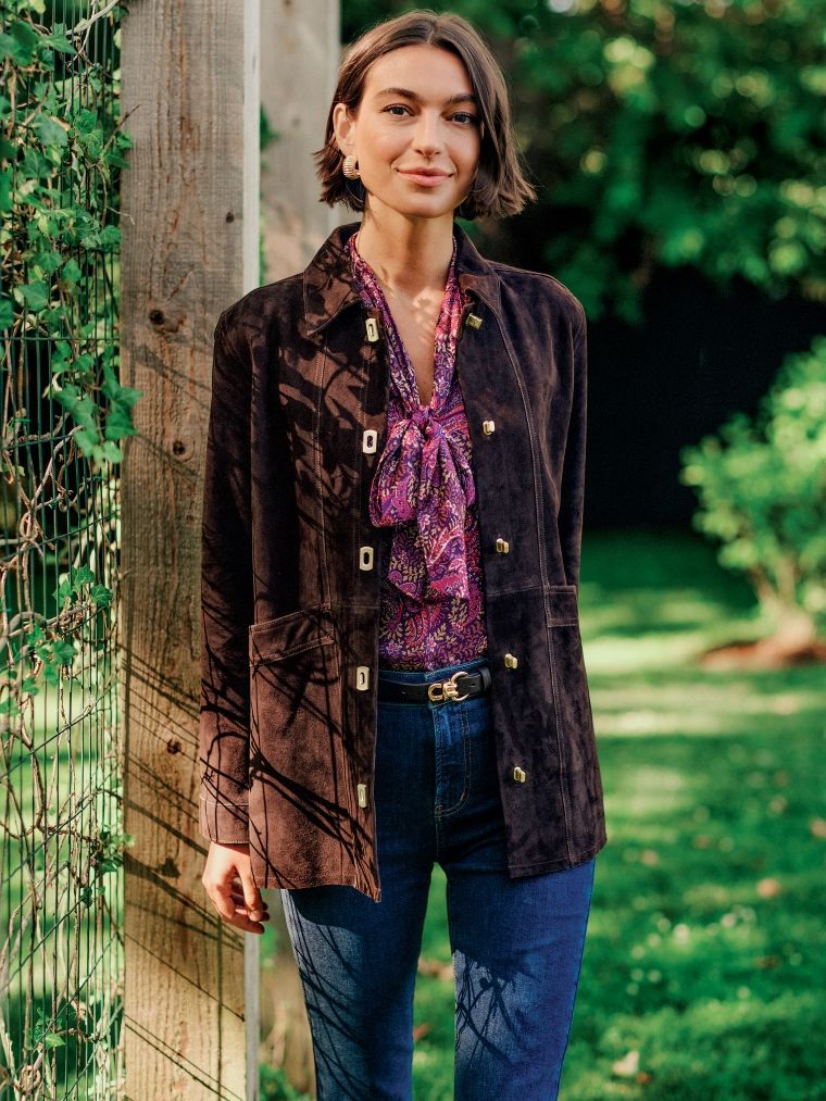Woman in a brown suede jacket, patterned purple blouse, and jeans stands next to a wooden post with ivy.