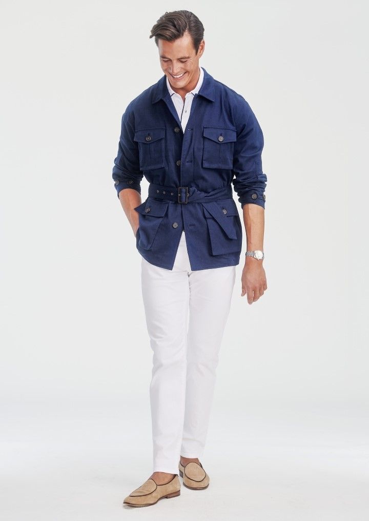 A man wearing a blue belted safari jacket over a white shirt, white pants, and tan loafers.