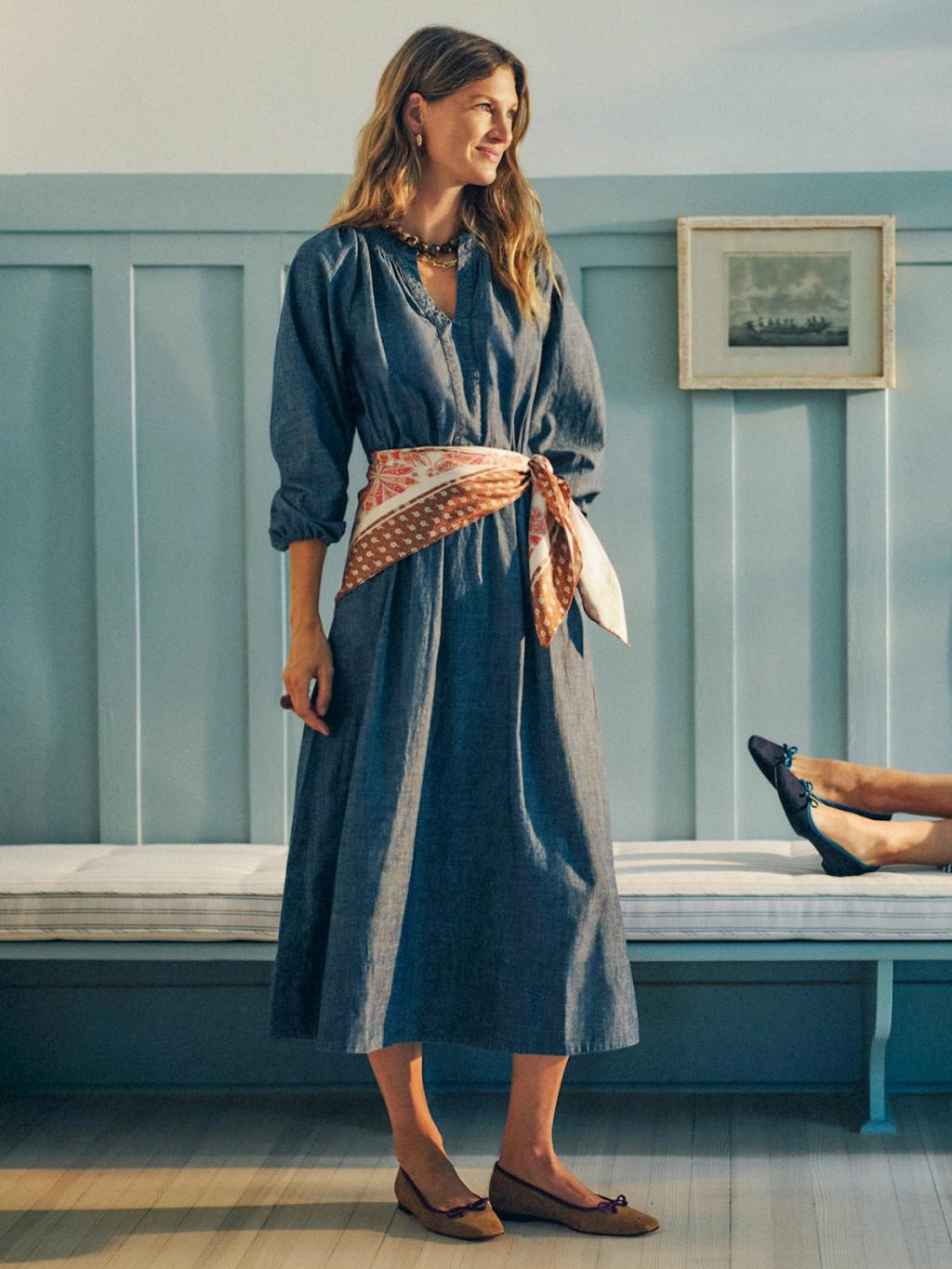 A woman in a blue chambray dress with a patterned sash stands beside a bench, near another person's feet.