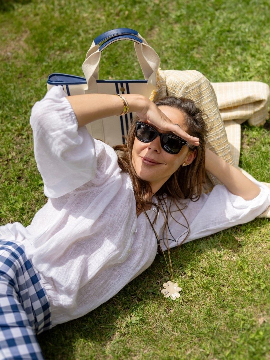 A woman wearing sunglasses lies on her side on green grass, shielding her eyes from the sun, with a tote bag and blanket behind her.