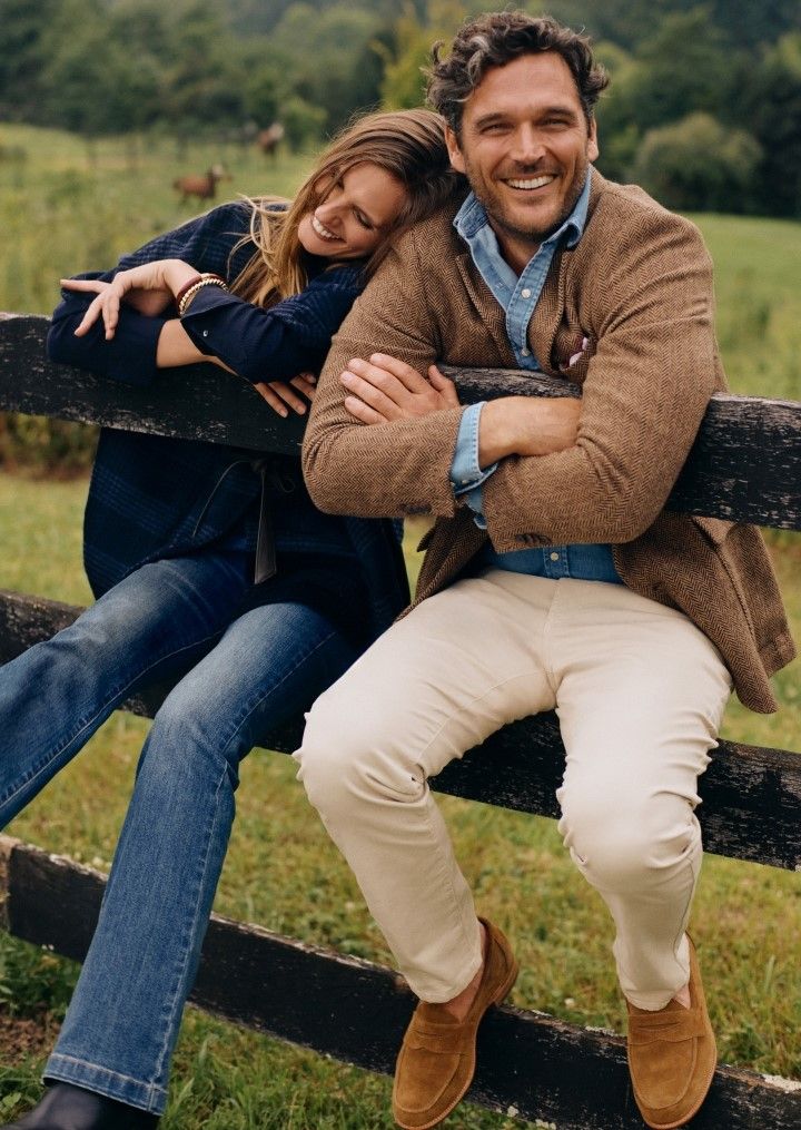 man and woman outside sitting on a fence, her wearing jeans and a navy plaid cape, him wearing khaki chino pants and a herringbone sport coat