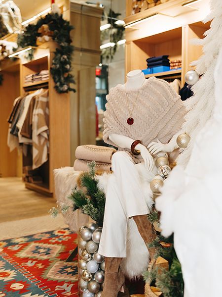 Indoor holiday retail display with a mannequin dressed in a textured beige poncho, white pants, and tan boots. The mannequin sits beside a white faux tree decorated with metallic ornaments and greenery. Shelves with neatly folded clothing and festive garlands decorate the store interior in the background.