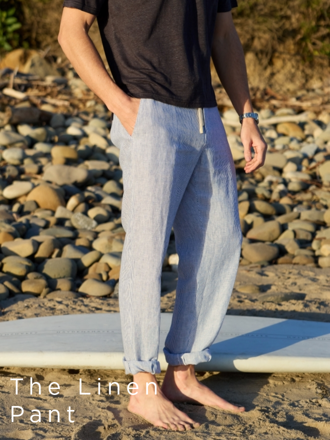 cropped image of bottom half of man on a beach wearing a navy knit shirt and blue hairline stripe linen pants rolled at the ankles