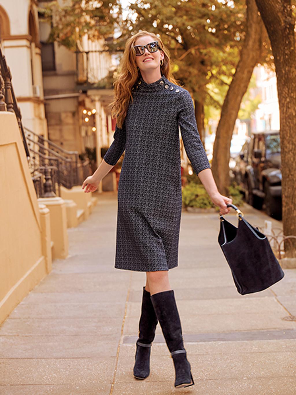 Women with long red hair in sunglasses walking down the street in a navy blue dress, tall black boots, and holding a black handbag.