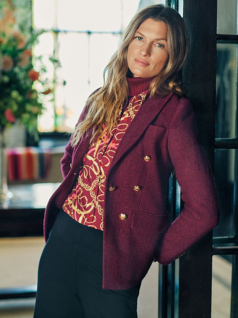 A woman with long wavy hair wears a burgundy blazer with gold buttons over a patterned blouse and a red turtleneck, leaning against a dark doorframe.