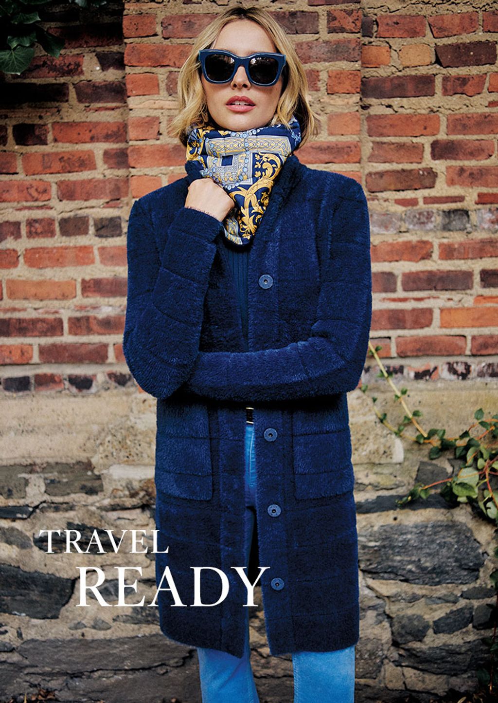 Woman wearing a navy coat, blue heraldic print snood, blue sunglasses and blue pants.