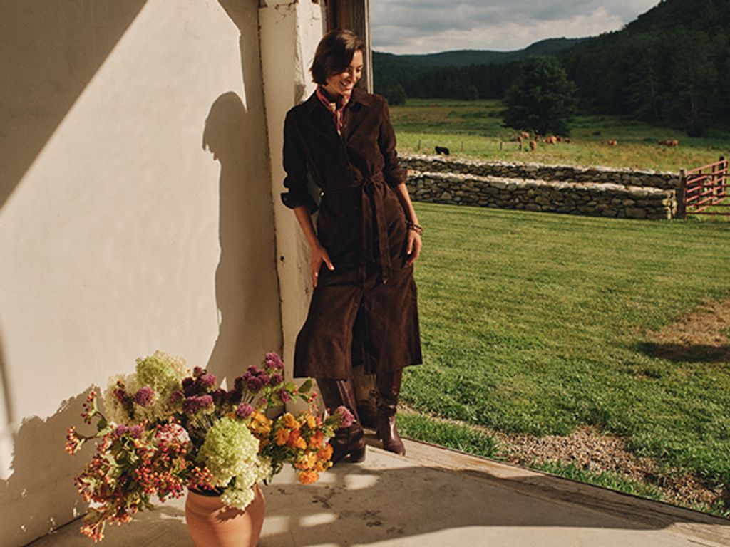 A woman in a dark brown belted coat leans in a doorway, with a vase of flowers in the foreground and a mountain landscape with cows in the background.