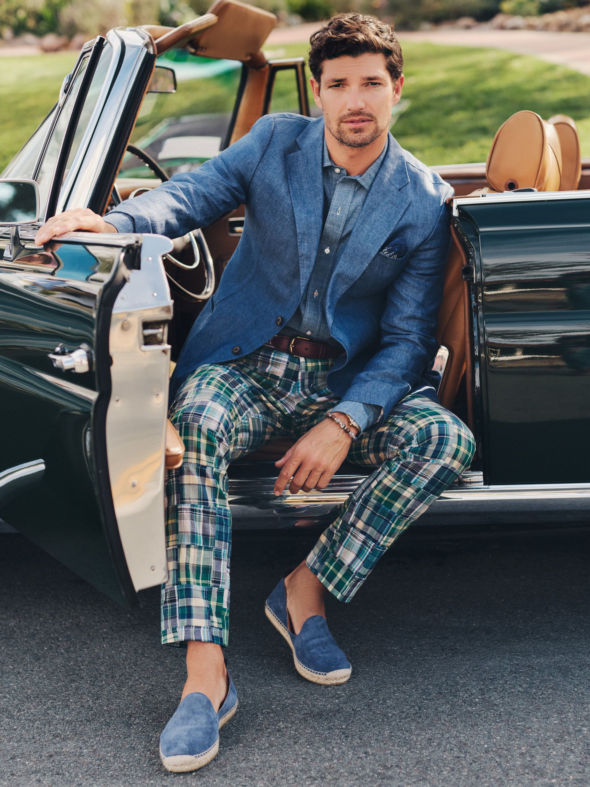 A man in a blue linen blazer, green plaid pants, and blue espadrilles sits in the doorway of a dark green convertible.