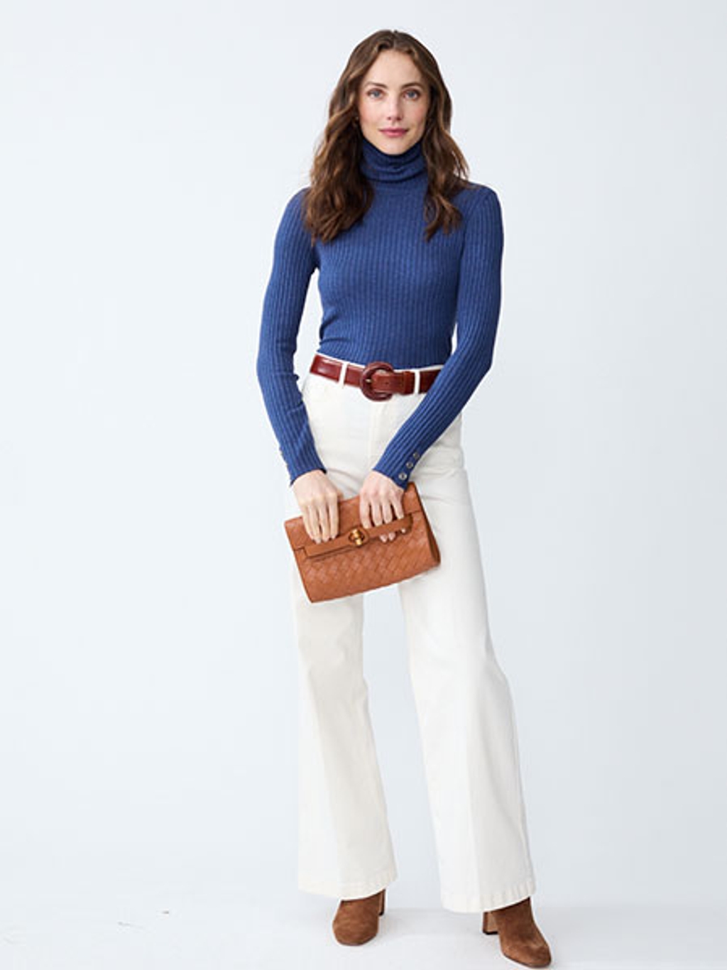 Women with brown hair wearing a navy blue turtleneck, white pants, holding a brown leather clutch.
