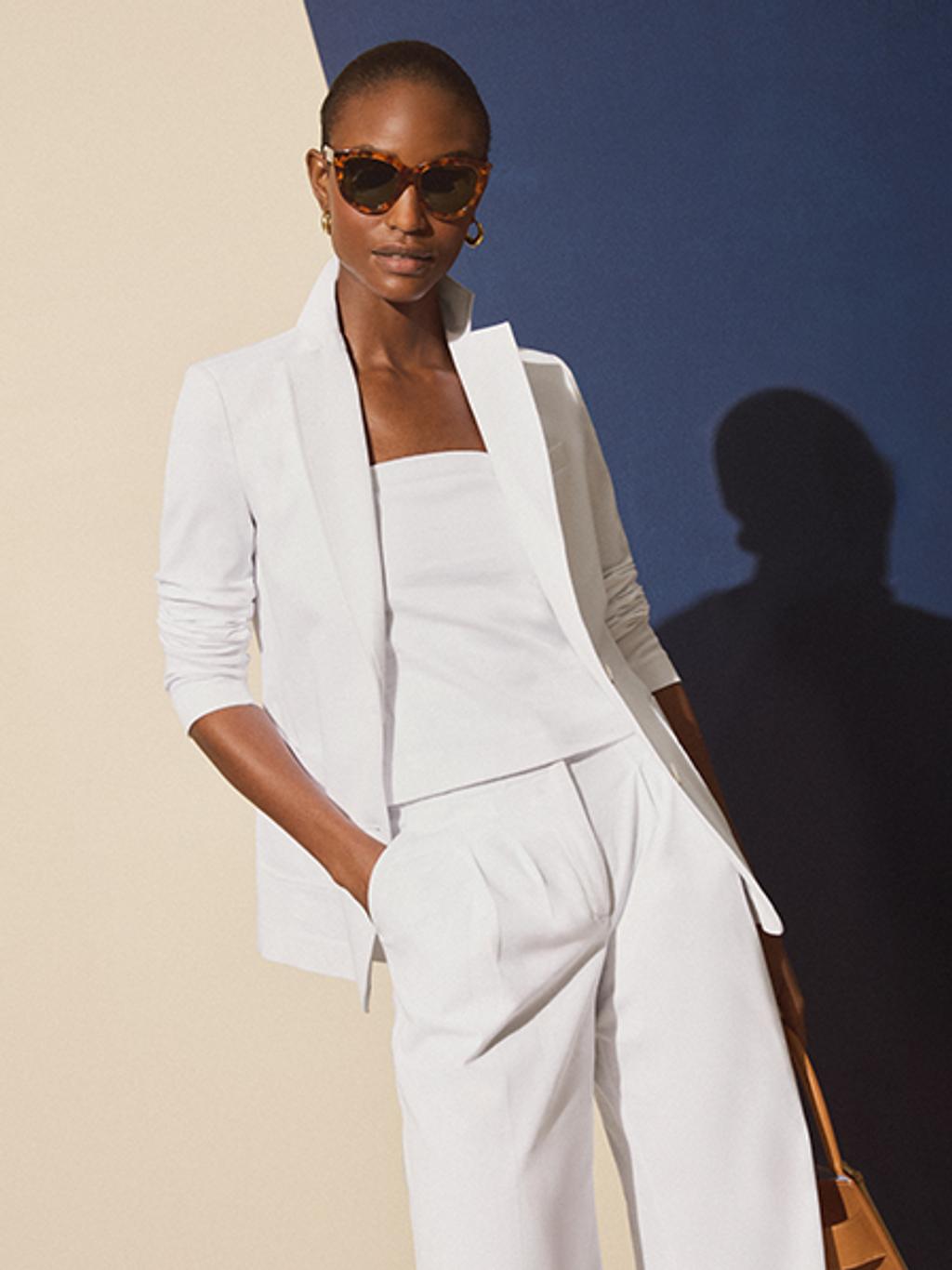 Model in tortoise sunglasses wearing a matching white suit with a white strapless tube top underneath. She is also holding a tan leather handbag.