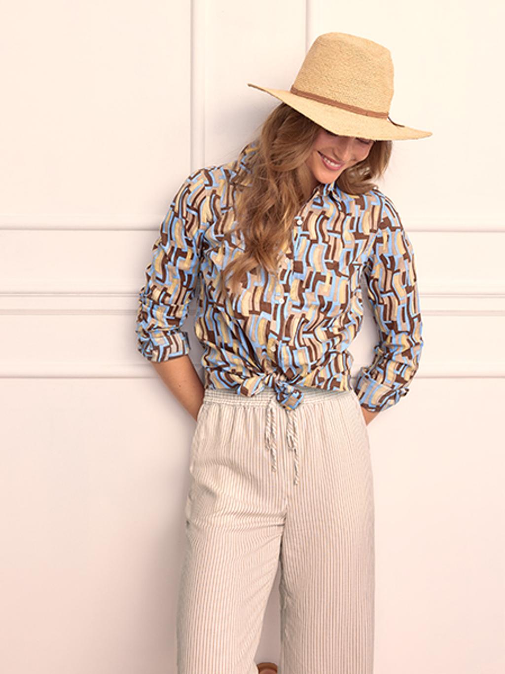 Woman leaning with her back against the wall, wearing a tan hat, patterned button up shirt, and striped pants.