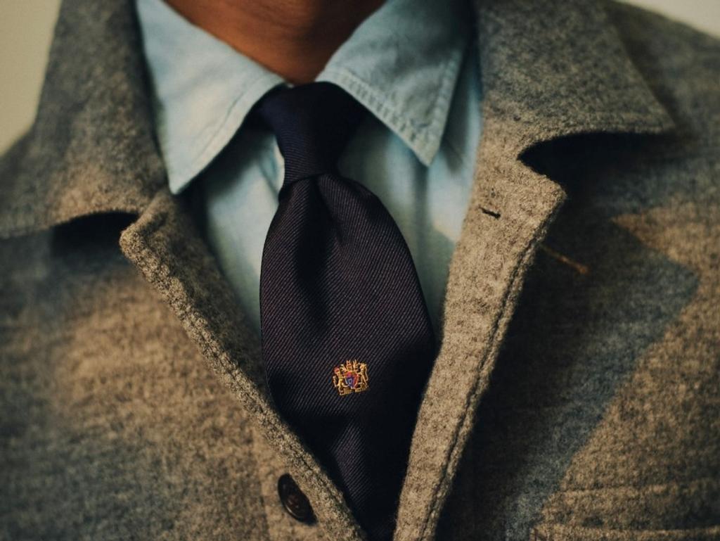 Close-up of a person wearing a grey woolen jacket, light blue shirt, and a dark blue tie with a small crest.