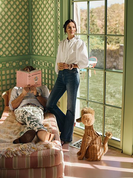 Female model in white dress shirt and jeans with gold wrist bangles posing next to male model laying on lounge with dress shirt and printed pants.
