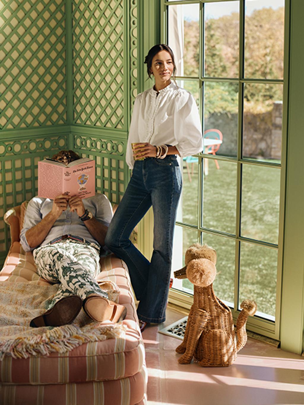 Female model in white dress shirt and jeans with gold wrist bangles posing next to male model laying on lounge with dress shirt and printed pants.