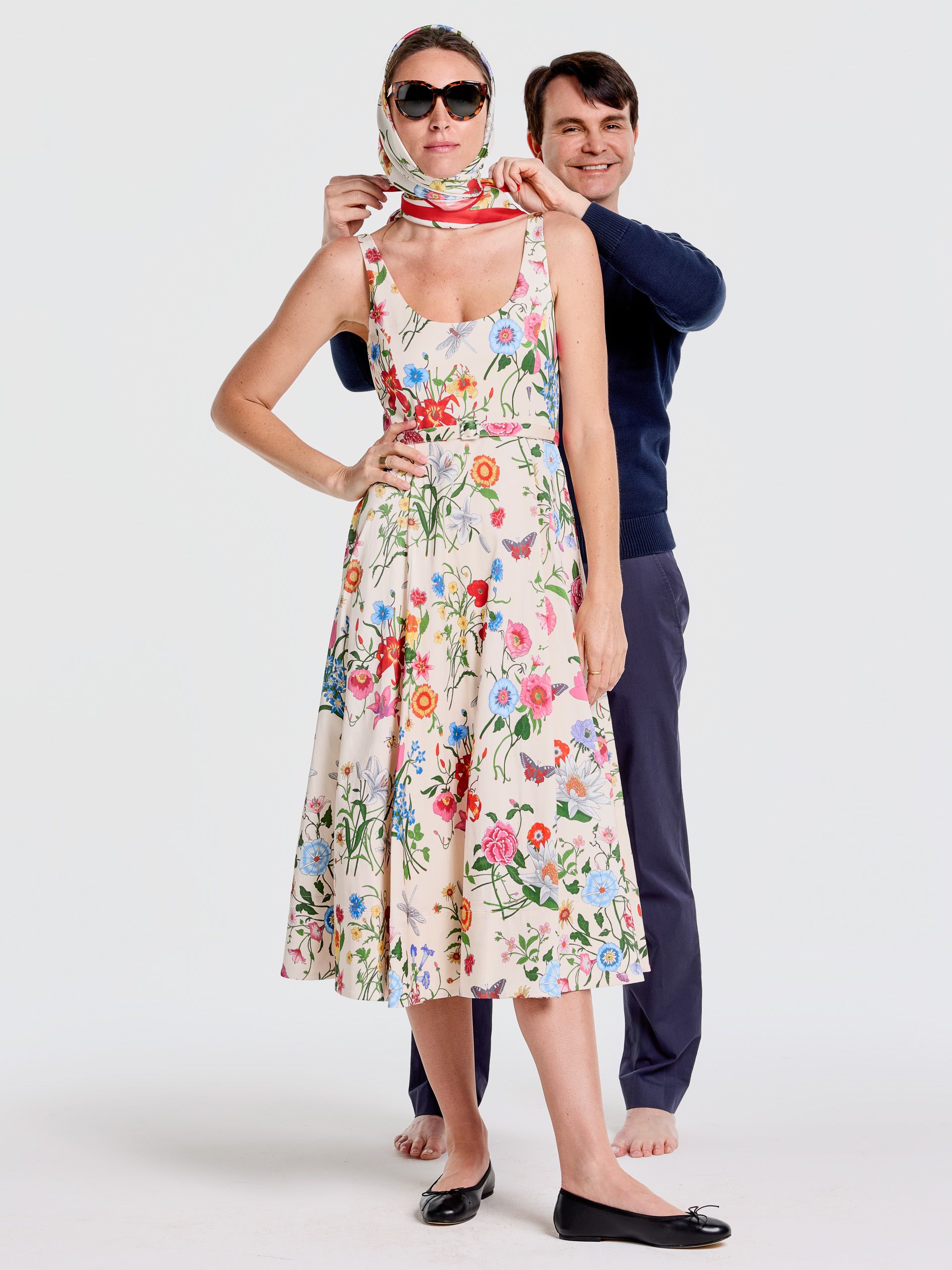 A man stands behind a woman in a floral dress and sunglasses, adjusting her patterned headscarf.