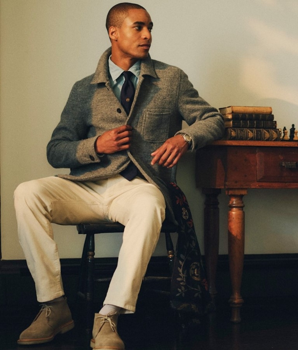 A man in a grey jacket, blue shirt, and cream pants sits on a stool. A desk with books and a chess set is to his right.