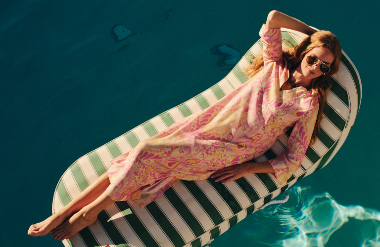A woman in a patterned pink dress and sunglasses floats on a striped pool raft.