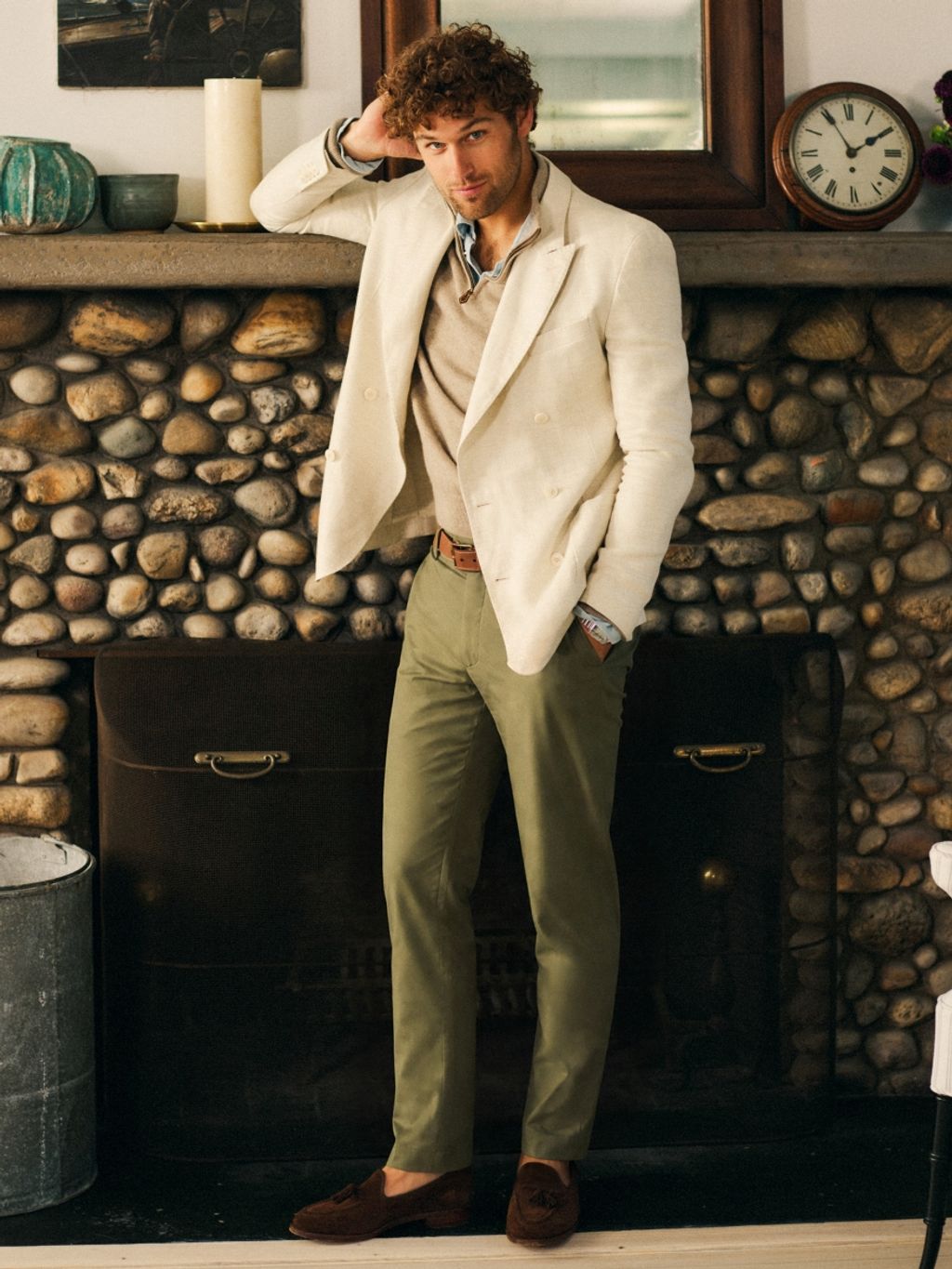 A man with curly hair, wearing a cream double-breasted blazer, beige sweater, and olive trousers, leans on a stone fireplace mantel.