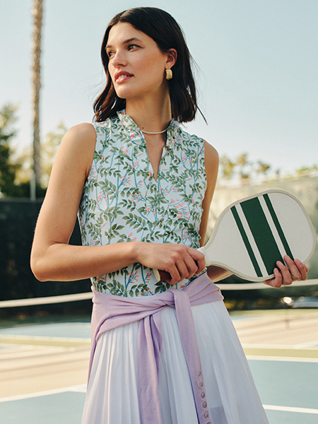 A woman in a floral top and white pleated skirt, with a purple sweater tied at her waist, holds a pickleball paddle on a court.