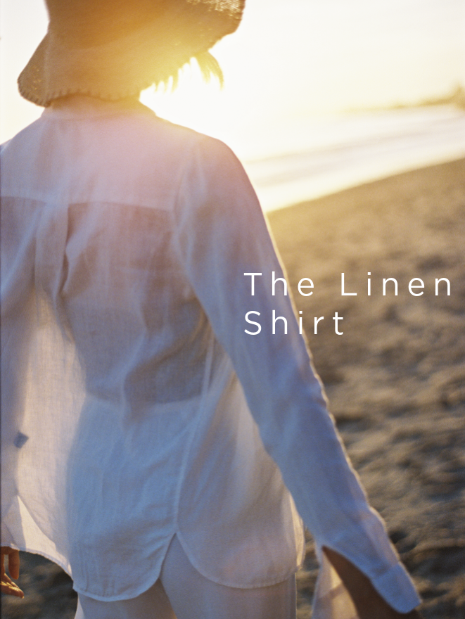 back of woman on the beach at sunset, wearing a white linen button down shirt and white linen pants with a straw hat